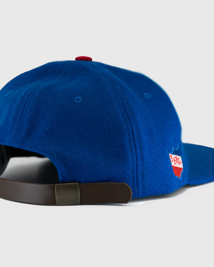 The EFF Milwaukee Clarks Vintage Ballcap is a blue wool cap with a brown adjustable strap and a red-and-white patch on the side, styled like a classic goalkeeper hat. Shown from the rear on a white background.