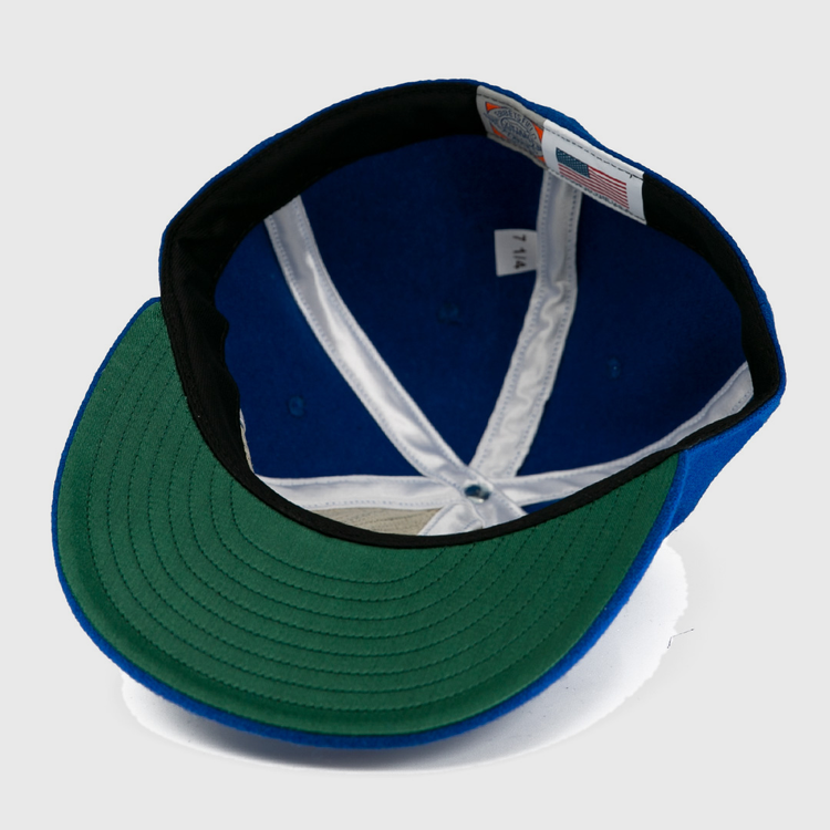 A close-up of the Milwaukee Clarks Vintage Ballcap by EFF shows a blue crown, green underbrim, white lining, black sweatband, and sewn size and flag tags—echoing classic International Hockey League style.