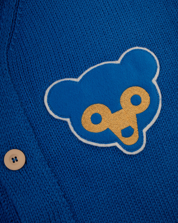 Close-up of the Ebbets Field Flannels Chicago Cubs 1962 Wool Shawl Collar Sweater, featuring a blue knit, beige button, and a blue and yellow stylized bear face patch.