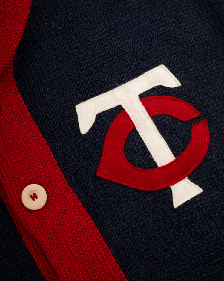 Close-up of a navy blue knit Minnesota Twins 1961-71 Wool Shawl Collar Sweater by Ebbets Field Flannels, featuring a white and red TC logo and a red buttoned edge with a white button, inspired by classic MLB field sweaters.