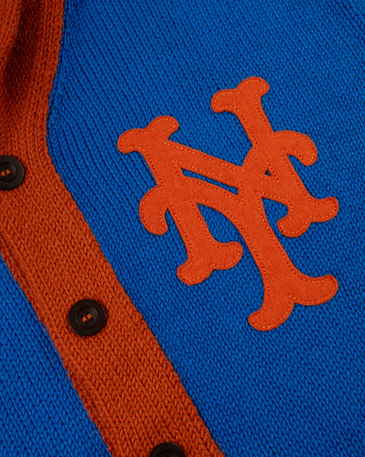 A close-up of the New York Mets 1962 Wool Shawl Collar Sweater by Ebbets Field Flannels, featuring blue knit fabric, an orange “NY” logo, orange trim, and black buttons—replicating a classic Mets on-field look.