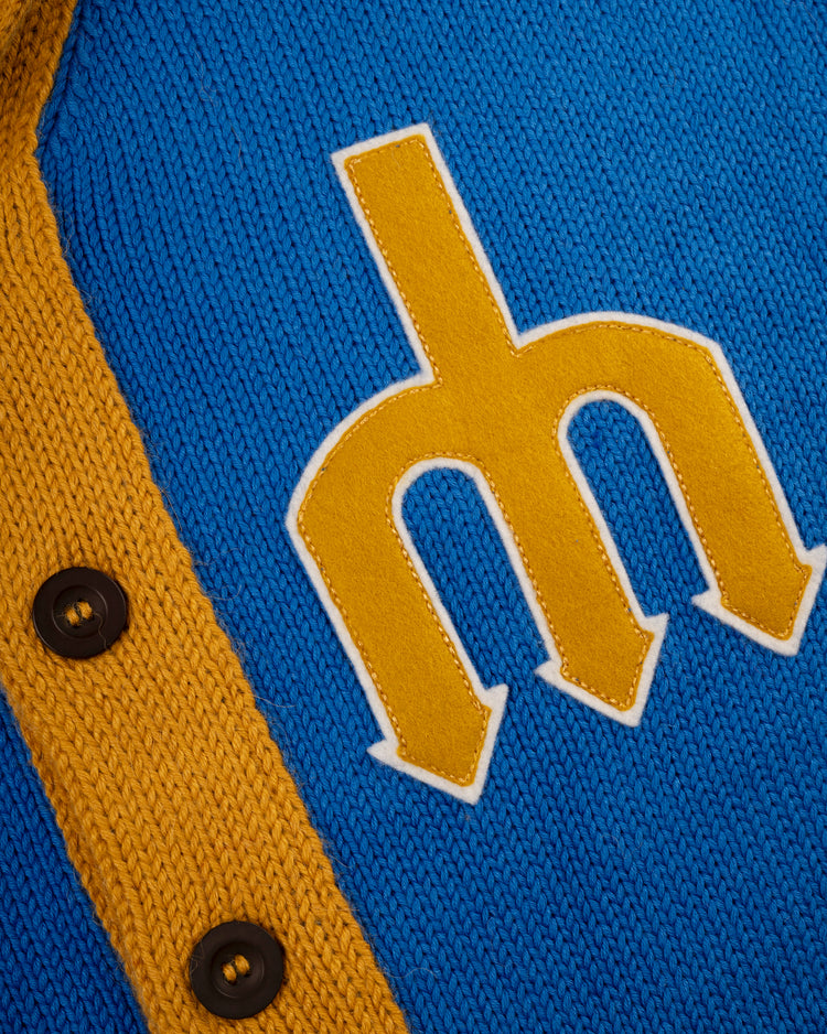 Close-up of the Seattle Mariners 1977-80 Wool Shawl Collar Sweater by Ebbets Field Flannels, featuring blue knit, a yellow stitched M, yellow trim, and two black buttons that highlight its classic replica MLB style.