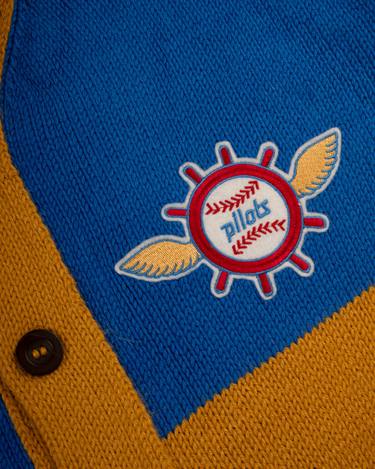 A close-up of the Ebbets Field Flannels Seattle Pilots 1969 Wool Shawl Collar Sweater, featuring blue and mustard yellow knit, a button, and a patch with a winged baseball and “Pilots” in blue.