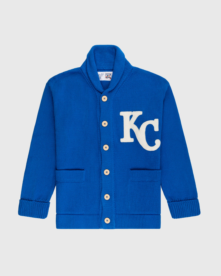 The Ebbets Field Flannels Kansas City Royals 1969 Wool Shawl Collar Sweater is a royal blue replica with tan buttons, two front pockets, a white KC patch on the left chest, and classic ribbed cuffs and hem.