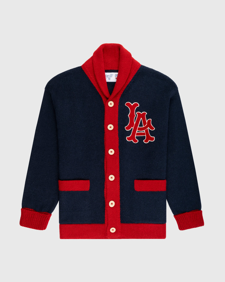 The Ebbets Field Flannels LA Angels 1961-65 Wool Shawl Collar Sweater is navy blue with red trim, cuffs, buttons, and pockets. Large red and white LA letters are embroidered on the left chest, inspired by vintage on-field sweaters.