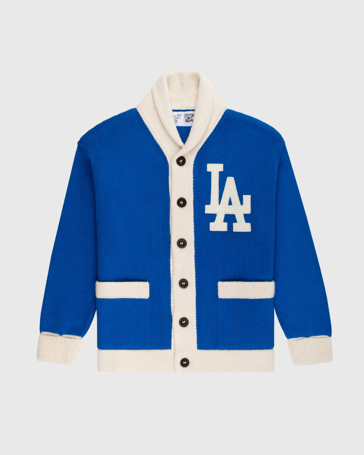 The Ebbets Field Flannels LA Dodgers 1958 Wool Shawl Collar Sweater is a blue and white varsity-style cardigan with black buttons, two front pockets, and a large LA logo on the left chest—replicating classic on-field baseball sweaters.