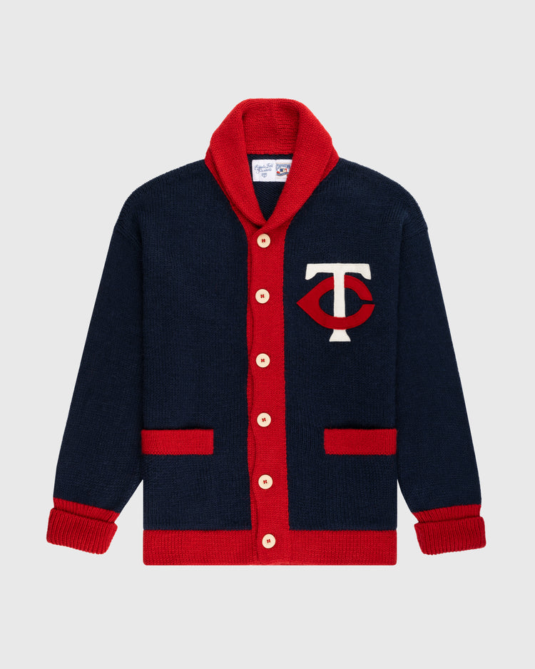 The Ebbets Field Flannels Minnesota Twins 1961-71 Wool Shawl Collar Sweater features navy wool, red trim, cream buttons, two red pockets, and a white T with a red eye-shaped design—perfect for true Twins fans seeking vintage style.