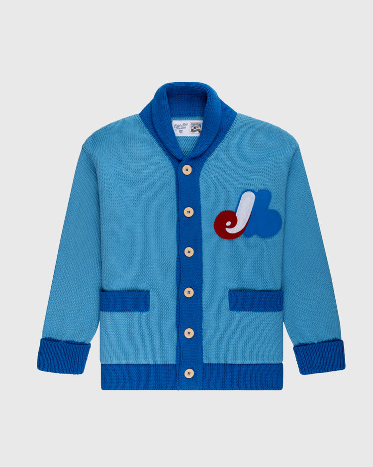 The Montreal Expos 1969 Wool Shawl Collar Sweater by Ebbets Field Flannels is a blue cardigan with darker blue trim, beige buttons, two front pockets, and a red, white, and blue chest patch inspired by classic baseball sweaters.