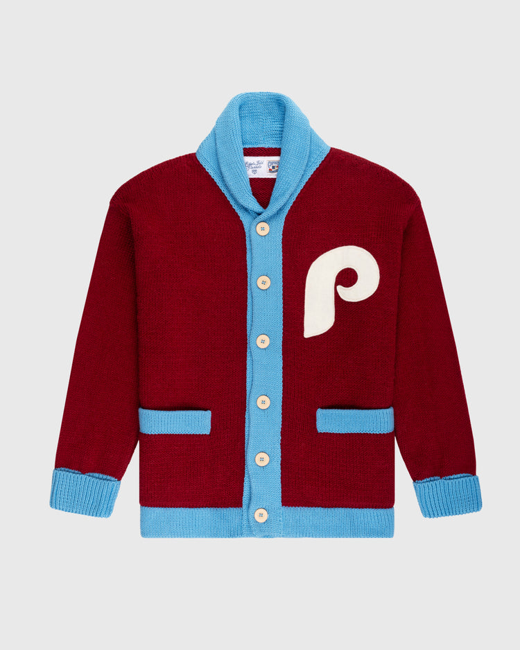 The Philadelphia Phillies 1979 Wool Shawl Collar Sweater by Ebbets Field Flannels features maroon wool, blue trim, cream buttons, blue cuffs and pockets, plus a large cream P on the chest—replicating classic on-field baseball sweaters. Light gray background.
