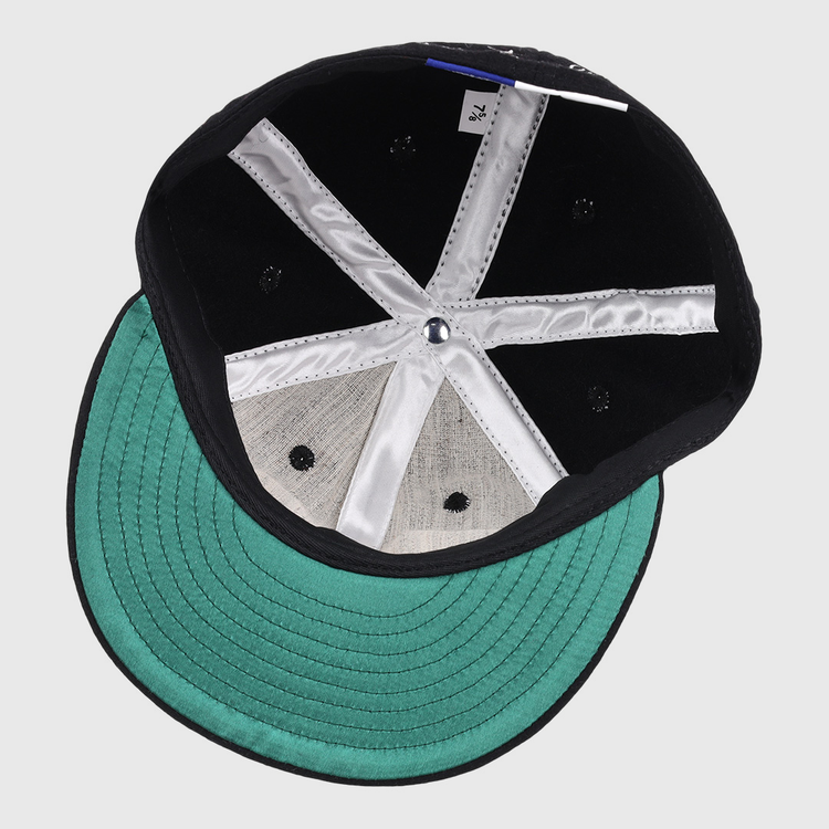 The image displays the inside of an Ebbets Field Flannels Monte Irvin Signature Series Ballcap, featuring a green underbrim, silver fabric lining, black rivets in the crown, and a white size tag—evoking early Negro National League styles.