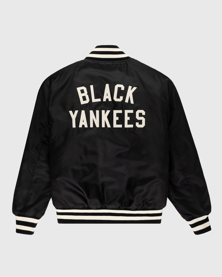 The Ebbets Field New York Black Yankees Satin Varsity Jacket is black with white striped cuffs, collar, and hem. The back displays large white BLACK YANKEES lettering, inspired by the NY Black Yankees. Shown on a plain light gray background.