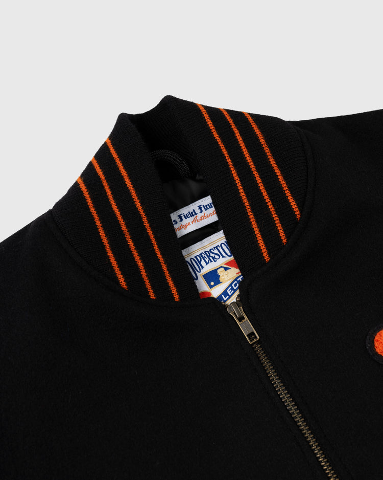 Close-up of the EFF New York Giants 1951 Authentic Wool & Leather Varsity Jacket in black, featuring a zip-up front, ribbed collar with orange stripes, and visible interior brand tags. The background is plain light gray.