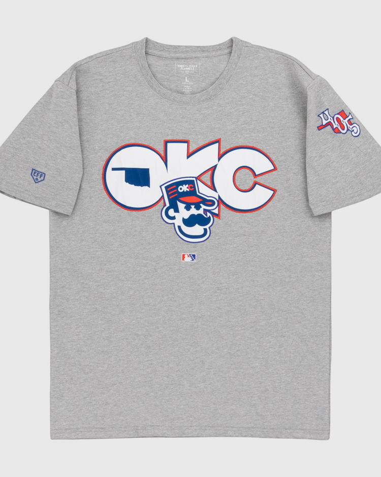 The Ebbets Field Oklahoma City 89Ers MiLB Fashion Cotton Short Sleeve Tee is a heather gray T-shirt with a large logo and cartoon character on the chest, plus 405 patches and other symbols on the sleeves.