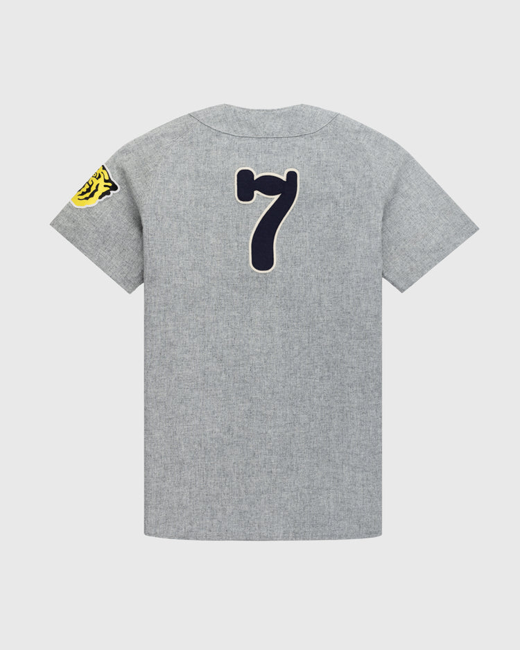 The Ebbets Field Flannels Osaka Tigers 1959 Authentic Wool Road Baseball Jersey features gray short sleeves, a large dark blue number 7 on the back, and a yellow tiger patch on the left sleeve, displayed on a plain white background.