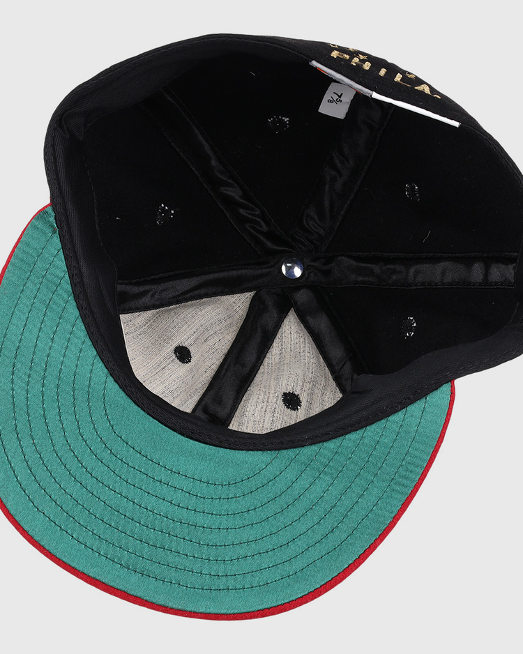 The EFF Philadelphia Stars Gilded Collection Ballcap is flipped upside down to reveal its green underside brim with red trim, black interior panels, blue center button, and a visible size tag—ideal for any Negro National League collector.