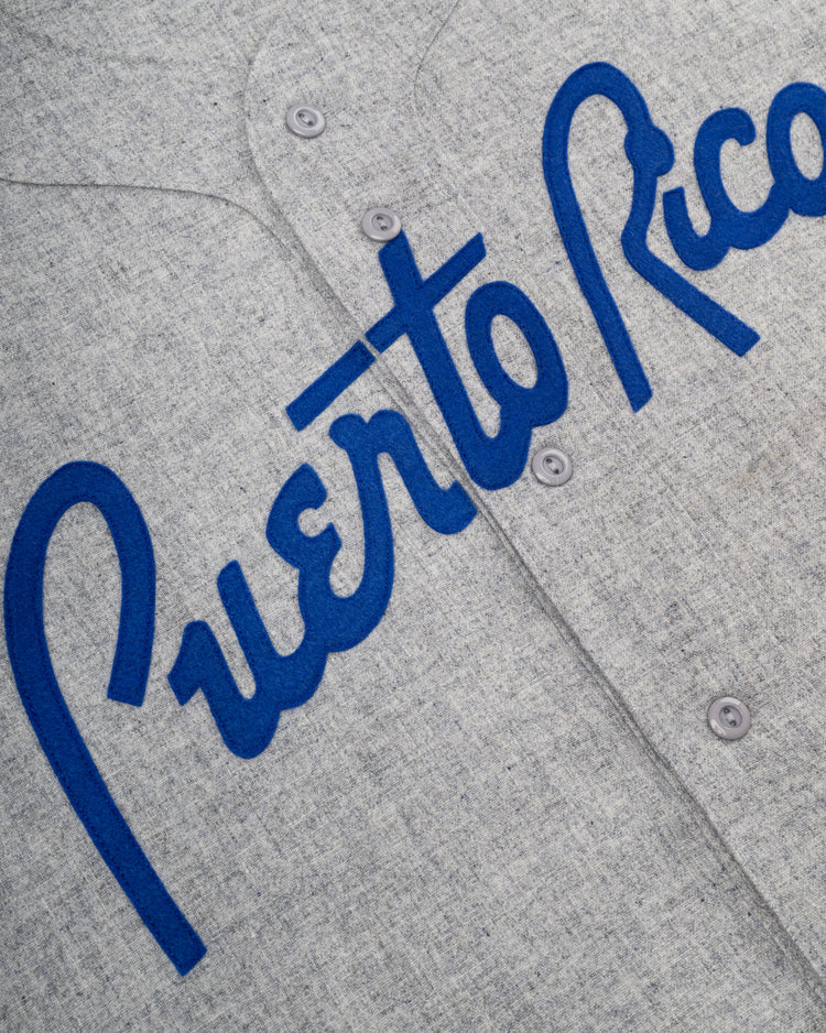 The Ebbets Field Puerto Rico 1958 National Team Authentic Wool Road Baseball Jersey features gray fabric, blue cursive Puerto Rico across the front, and visible buttons, inspired by the original team jersey.