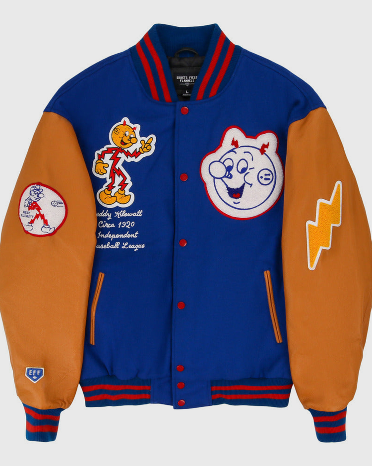 The Ebbets Field Reddy Kilowatt DNA Varsity Jacket is blue and orange, with cartoon baseball patches, smiling baseball faces, a lightning bolt, and an “Officially Licensed Cooperstown Baseball League” patch.