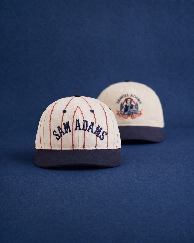 Two beige Sam Adams Pinstripe Wool Ballcaps by Ebbets Field with navy brims rest on a blue background. The front displays embroidered SAM ADAMS; the back features a Samuel Adams graphic. A unique spin on the classic Ebbets Field cap.