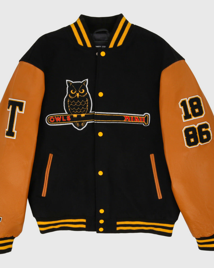 The Ebbets Field Topeka Owls MiLB Dna Varsity Jacket is black with tan leather sleeves, features an owl-on-bat patch, OWLS text, 1886 on the right sleeve, a large T on the left sleeve, and yellow striped collar and cuffs.