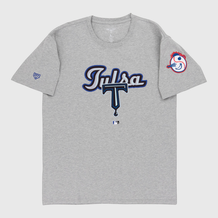 The Ebbets Field Tulsa Drillers MiLB Fashion Cotton Short Sleeve Tee is a gray shirt with “Tulsa” in blue and white script and a large T on the front. The left sleeve features the Drillers’ baseball mascot head with a red feather headband.