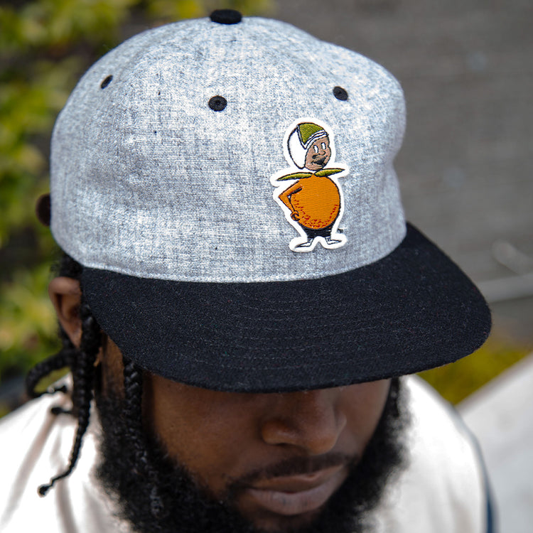 A man wears the EFF University of Central Florida Mascot Vintage Ballcap, a grey and black hat featuring a cartoon orange mascot with arms, legs, a face, and a green cap, standing out against the blurred greenery behind him.