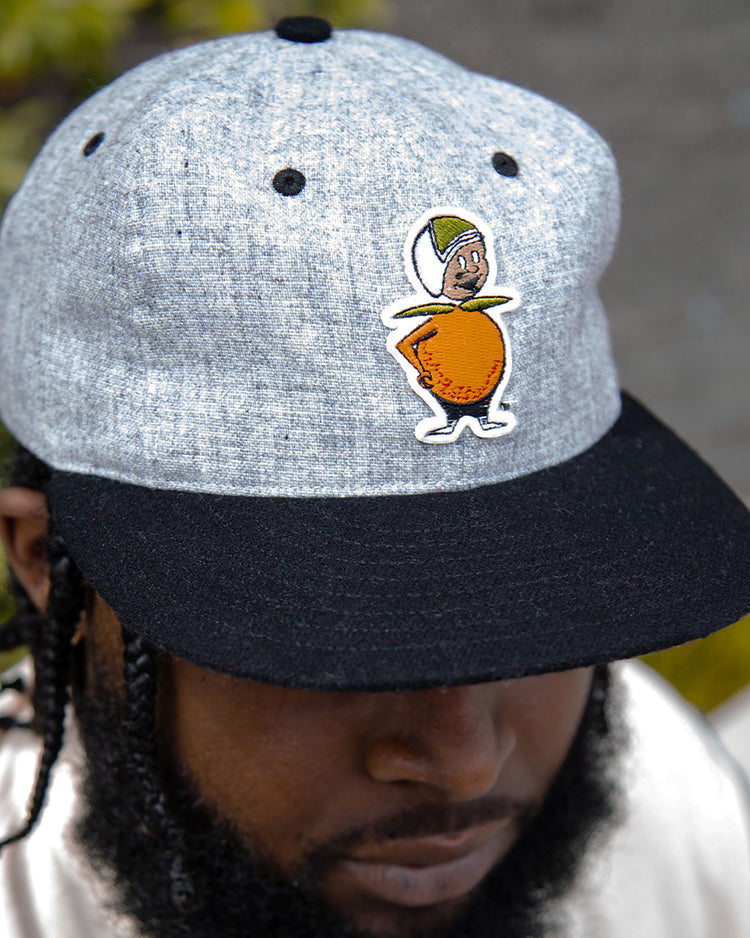 A man wears the EFF University of Central Florida Mascot Vintage Ballcap, a grey and black hat featuring a cartoon orange mascot with arms, legs, a face, and a green cap, standing out against the blurred greenery behind him.