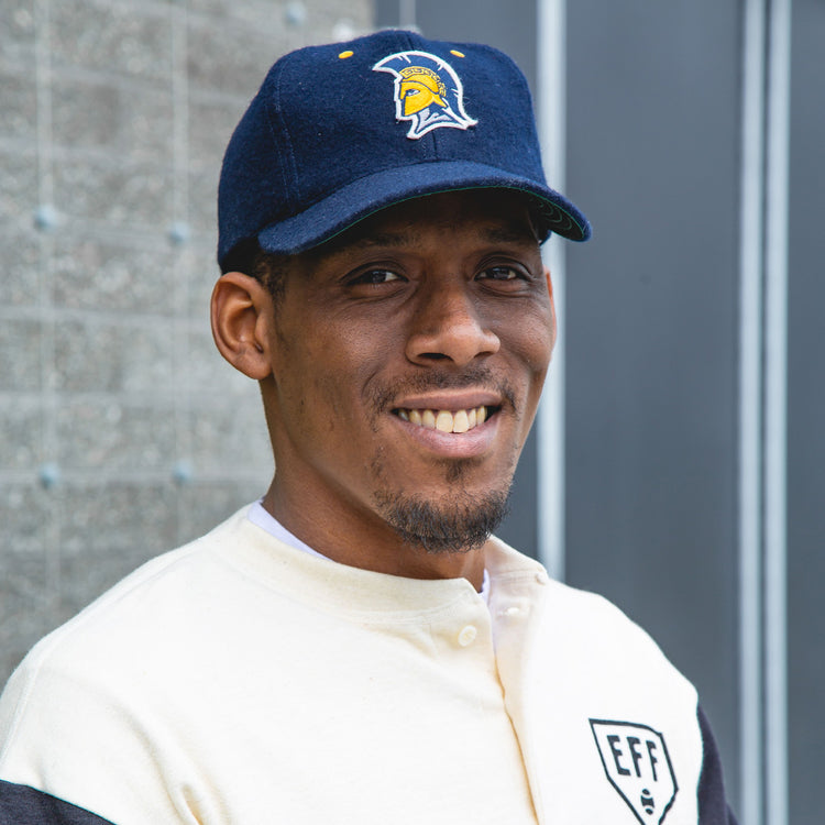 A man wearing the EFF University of North Carolina Greensboro Mascot Vintage Ballcap with a yellow Spartan helmet logo and a cream and black jacket smiles outdoors in front of a textured wall.
