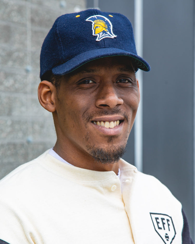 A man wearing the EFF University of North Carolina Greensboro Mascot Vintage Ballcap with a yellow Spartan helmet logo and a cream and black jacket smiles outdoors in front of a textured wall.