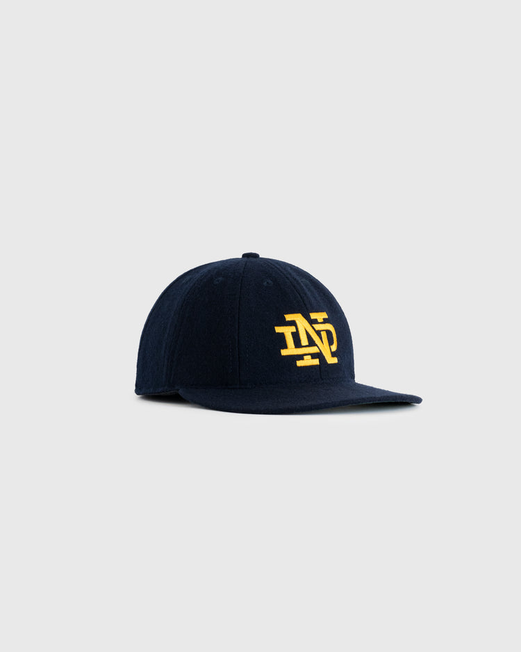 The EFF University of Notre Dame 1943 Authentic Wool 8-Panel Ballcap is a navy vintage cap with a flat brim, featuring a yellow intertwined ND logo—an homage to the 43 football champs—shown on a plain light gray background.