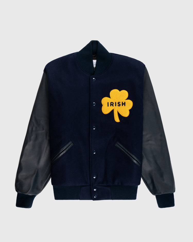 The EFF University of Notre Dame Authentic Wool & Leather Varsity Jacket features a navy body, black leather sleeves, black ribbed cuffs and collar, and a large yellow shamrock patch with “IRISH” on the chest.