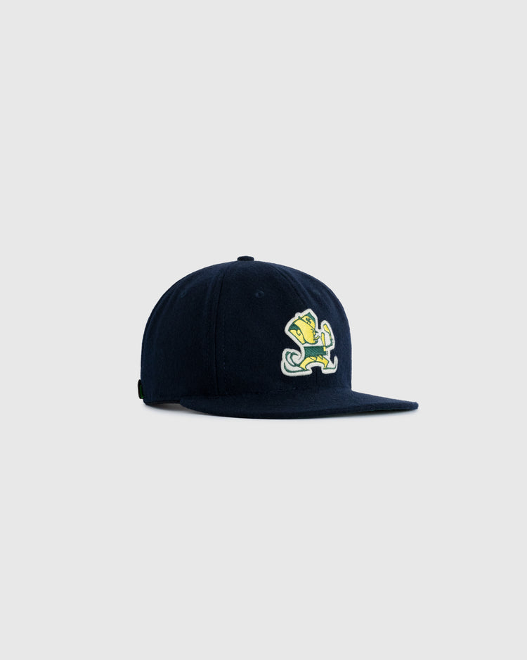 The EFF University of Notre Dame Leprechaun Authentic Wool Ballcap in navy features a cartoon mascot with a trophy on the front, set against a light gray background—a playful tribute to collegiate athletics.