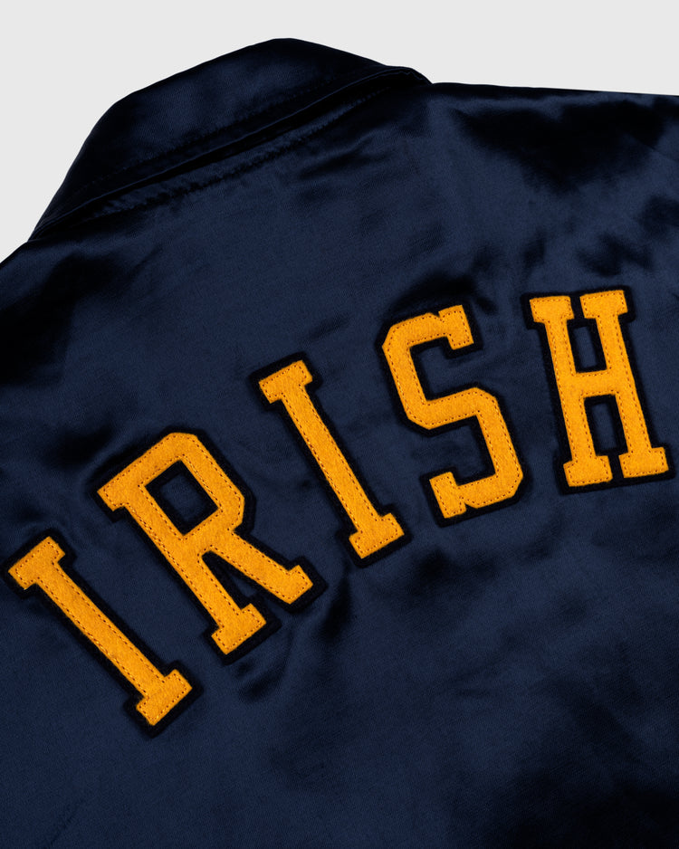 The EFF University of Notre Dame Satin Windbreaker Jacket features a vintage-inspired navy blue design with bold yellow IRISH lettering across the back, echoing the style of a classic coach’s jacket.