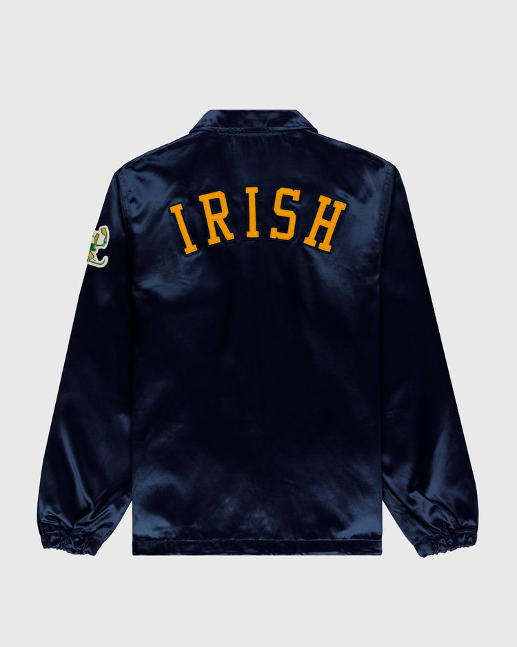 The EFF University of Notre Dame Satin Windbreaker Jacket is a vintage-inspired navy blue jacket with yellow IRISH lettering on the back and an embroidered patch on the left sleeve, shown against a plain light background.