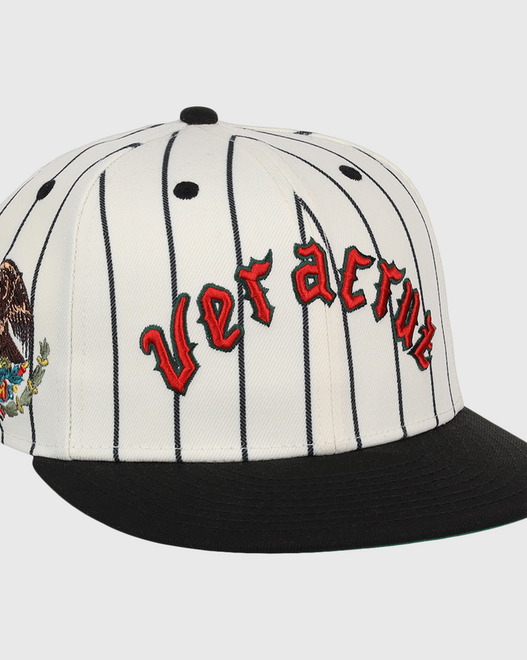 The Veracruz NLB Pinstripe Fitted Ballcap by EFF is a white cap with black pinstripes, a black brim, Veracruz in red gothic letters on the front, and an eagle-and-cactus embroidered patch on the side.