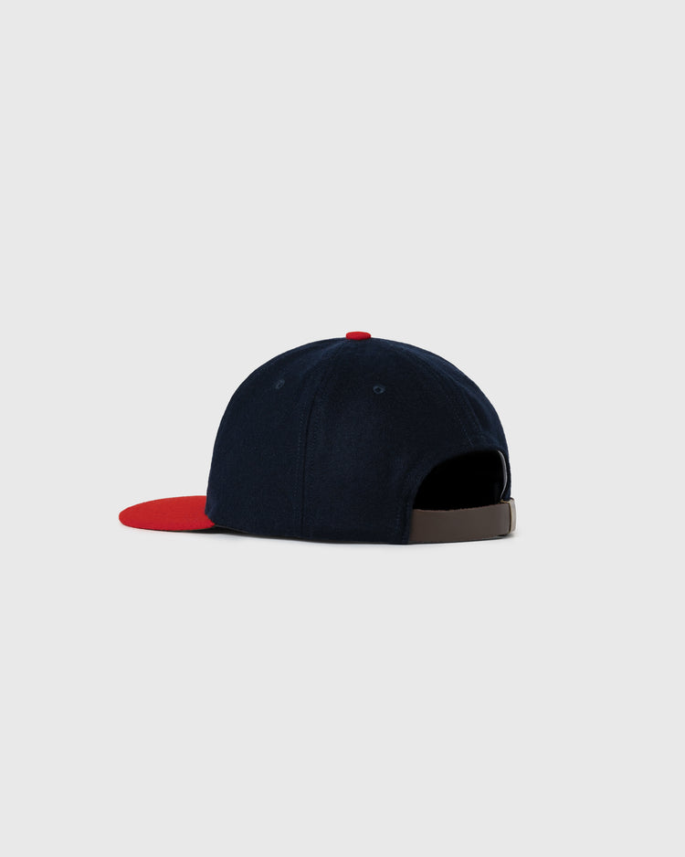 The Wichita Braves 1957 Authentic Wool Ballcap by Ebbets Field Flannels features navy blue wool, a red brim, and a brown adjustable strap, shown against a plain light gray background.