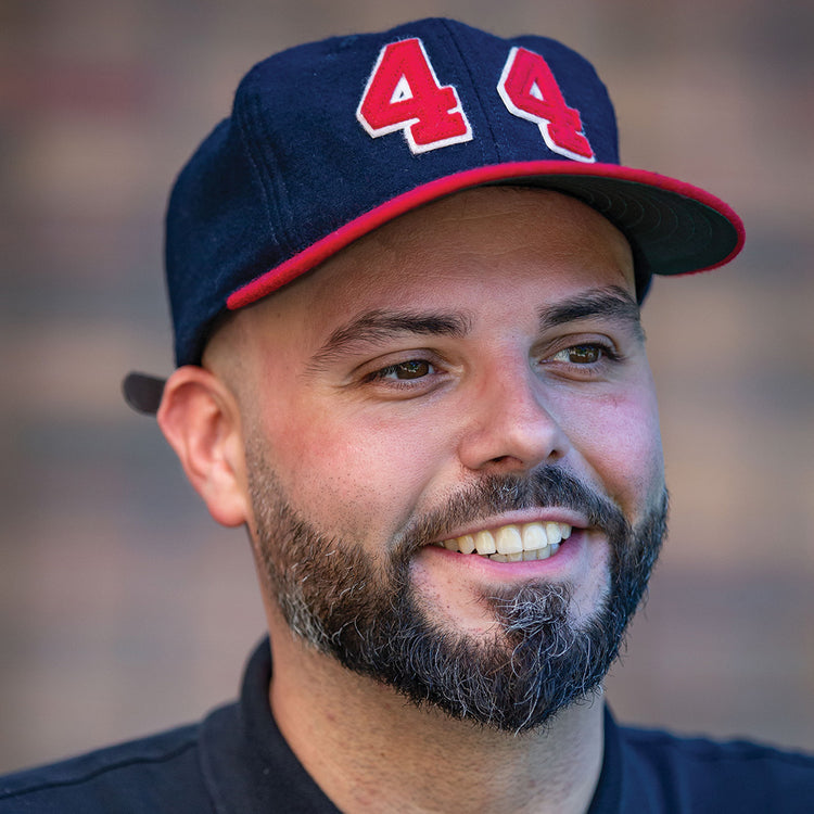 A bearded man smiles while wearing the Hank Aaron 1957 Signature Series Ballcap by Ebbets Field Flannels—a navy wool cap with red trim, the number 44, and Hank Aaron’s signature. The background is blurred.