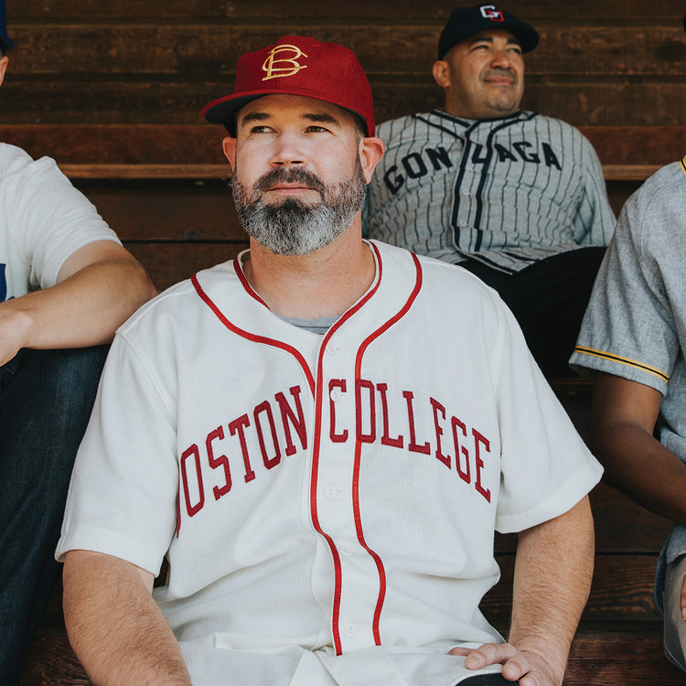 A man with a gray beard in an EFF Boston College 1949 Home Jersey and matching cap sits on wooden bleachers, surrounded by others wearing various vintage baseball jerseys.