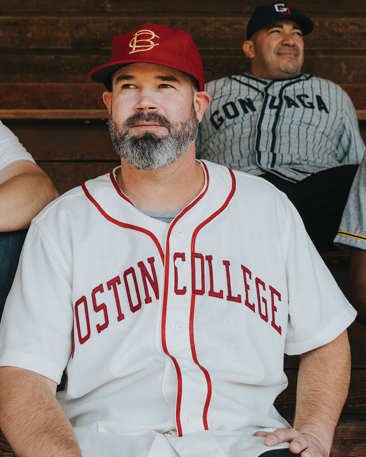 A man with a gray beard in an EFF Boston College 1949 Home Jersey and matching cap sits on wooden bleachers, surrounded by others wearing various vintage baseball jerseys.