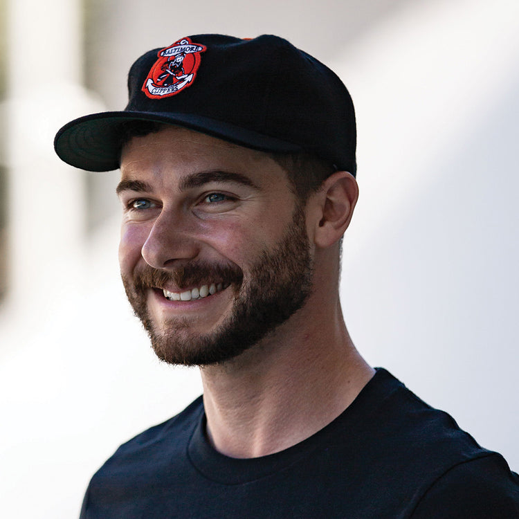 A smiling man with a short beard wears a black T-shirt and the EFF Baltimore Clippers 1962 Vintage Ballcap, featuring a red and white embroidered patch, as he stands against a blurred light background.