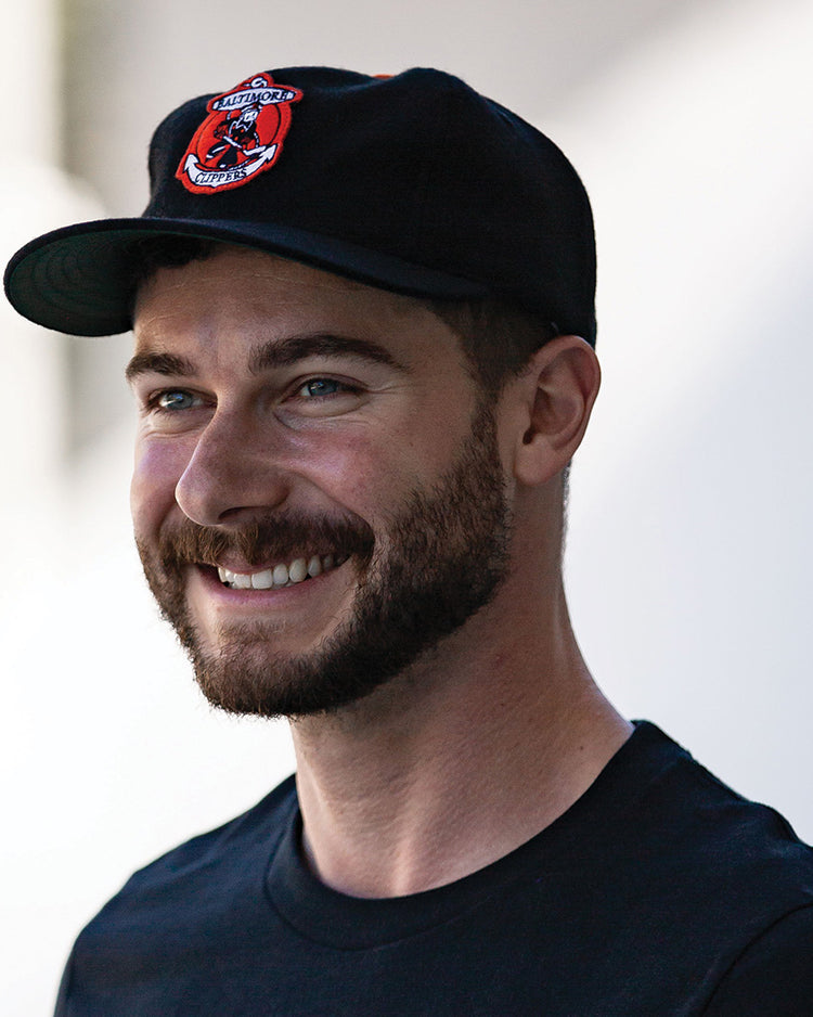 A smiling man with a short beard wears a black T-shirt and the EFF Baltimore Clippers 1962 Vintage Ballcap, featuring a red and white embroidered patch, as he stands against a blurred light background.