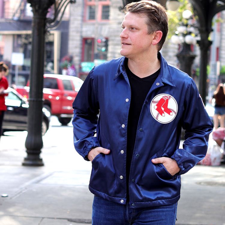 A man wearing an EFF Boston Red Sox 1967 Satin Windbreaker and jeans stands on a city street with one hand in his pocket, looking to the side. Cars and people are visible in the blurred background.