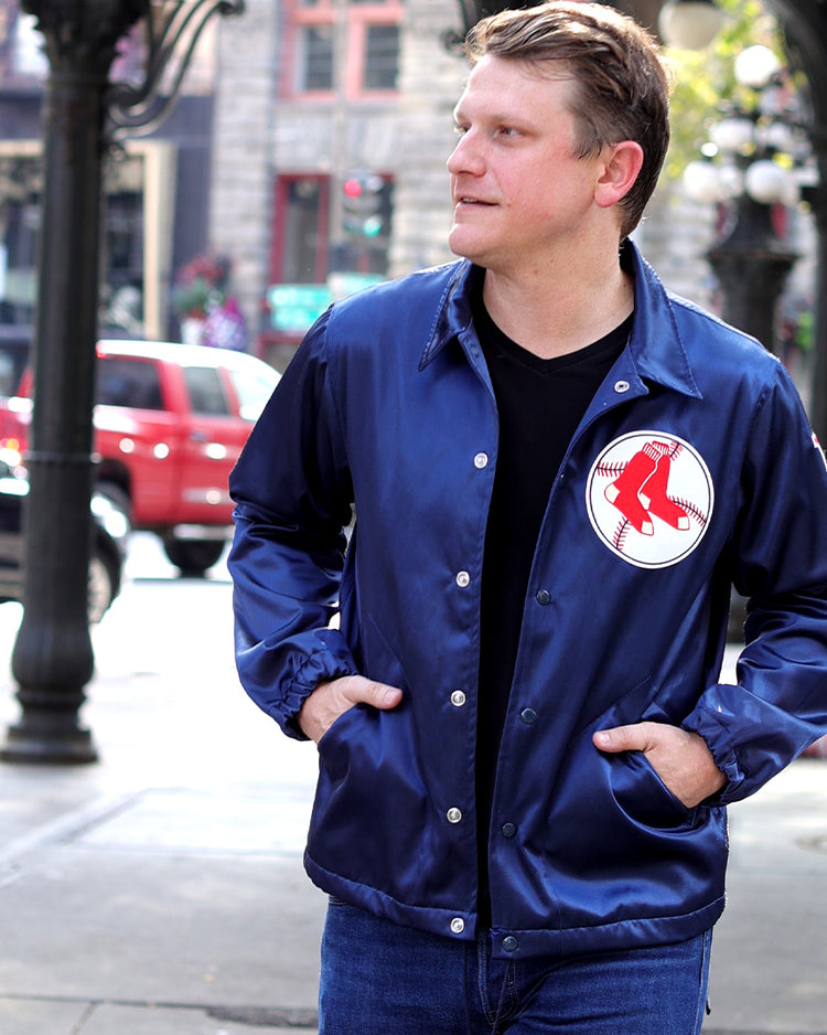 A man wearing an EFF Boston Red Sox 1967 Satin Windbreaker and jeans stands on a city street with one hand in his pocket, looking to the side. Cars and people are visible in the blurred background.