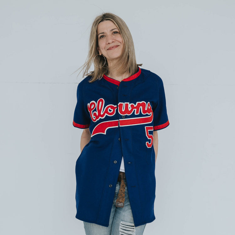 A smiling woman with blond hair wears an EFF Indianapolis Clowns 1952 Road Jersey, featuring Clowns in red and white letters and number 5, honoring the historic Negro Leagues team. She stands against a plain light background.