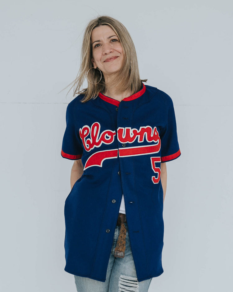 A smiling woman with blond hair wears an EFF Indianapolis Clowns 1952 Road Jersey, featuring Clowns in red and white letters and number 5, honoring the historic Negro Leagues team. She stands against a plain light background.