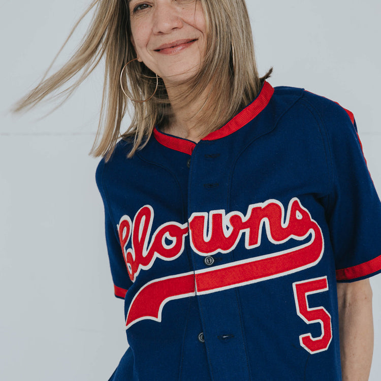 A smiling woman with straight, light brown hair wears an EFF Indianapolis Clowns 1952 Road Jersey in blue with red trim and “Clowns” script across the front. The number 5 references the legendary Negro Leagues team. Her face is partially out of frame.