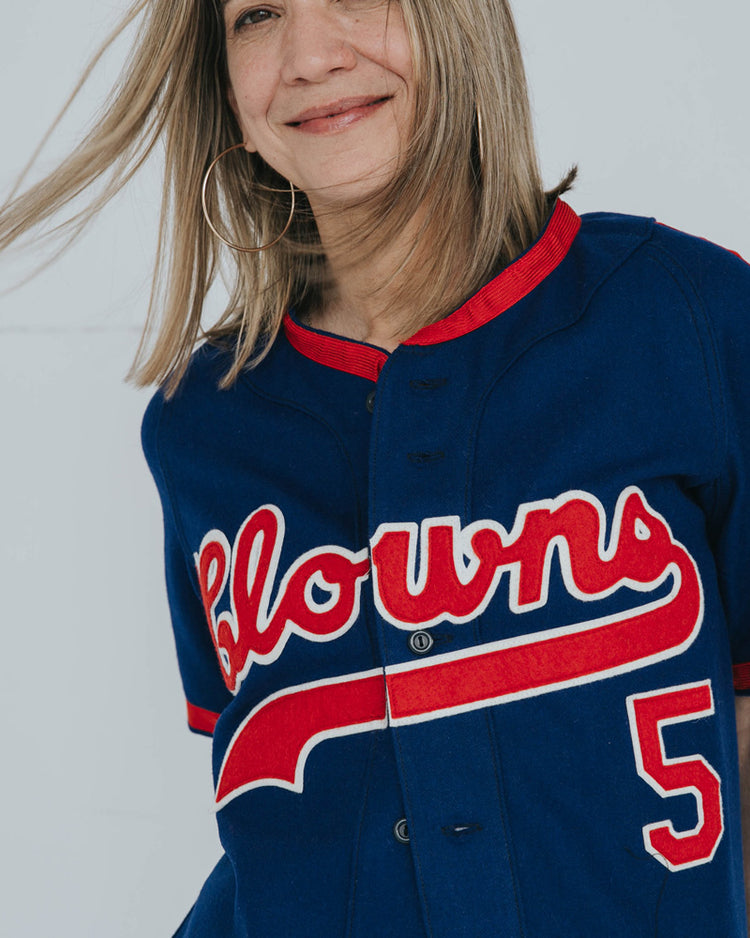 A smiling woman with straight, light brown hair wears an EFF Indianapolis Clowns 1952 Road Jersey in blue with red trim and “Clowns” script across the front. The number 5 references the legendary Negro Leagues team. Her face is partially out of frame.