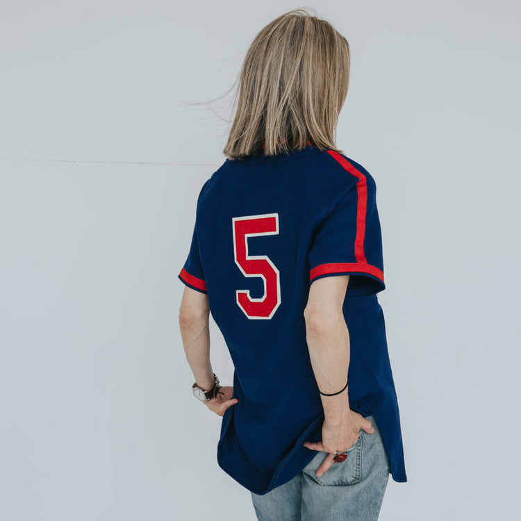 A person with shoulder-length blonde hair, wearing an EFF Indianapolis Clowns 1952 Road Jersey in blue/red with number 5, stands facing away with a phone in hand and light blue jeans.