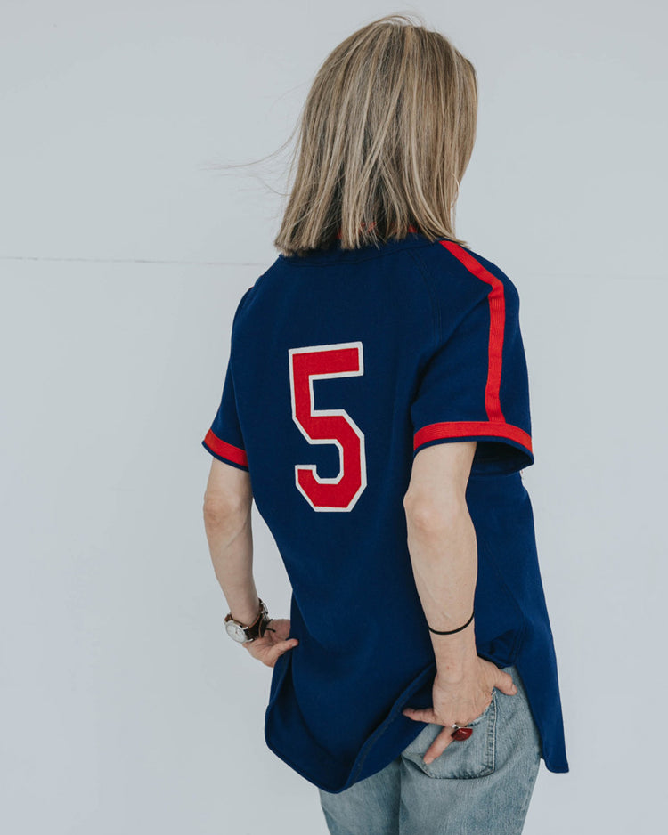 A person with shoulder-length blonde hair, wearing an EFF Indianapolis Clowns 1952 Road Jersey in blue/red with number 5, stands facing away with a phone in hand and light blue jeans.