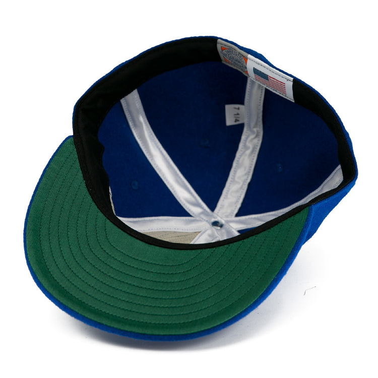 A close-up of the Milwaukee Clarks Vintage Ballcap by EFF shows a blue crown, green underbrim, white lining, black sweatband, and sewn size and flag tags—echoing classic International Hockey League style.