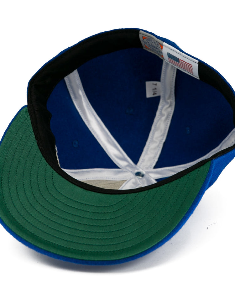 A close-up of the Milwaukee Clarks Vintage Ballcap by EFF shows a blue crown, green underbrim, white lining, black sweatband, and sewn size and flag tags—echoing classic International Hockey League style.